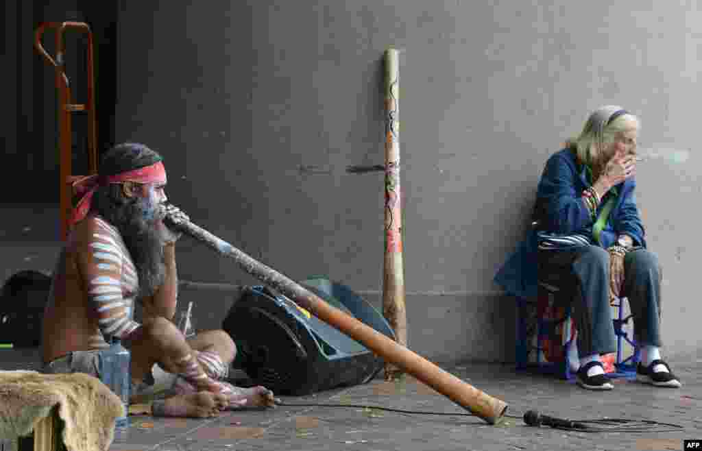 An elderly woman smokes a cigarette while an Aboriginal man plays a didgeridoo in Circular Quay in Sydney, Australia.