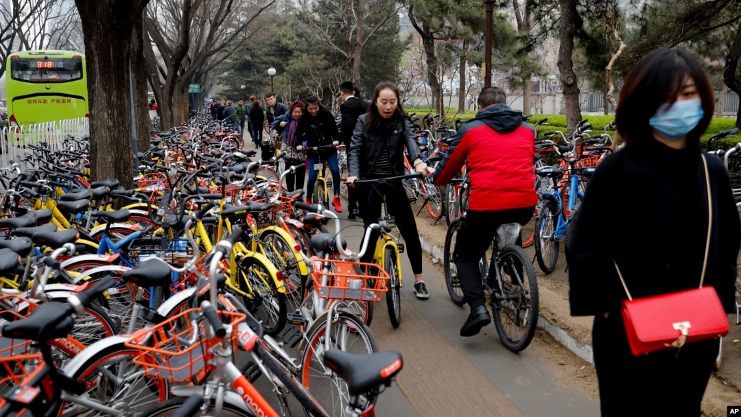 Residents ride bicycles from bike-sharing company Ofo try to pedal through a sidewalk crowded with bicycles from the bike-sharing companies Ofo, Mobike and Bluegogo, near a bus stand in Beijing, China, March 23, 2017.