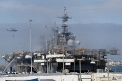 A helicopter carrying water passes the USS Bonhomme Richard, July 14, 2020, in San Diego. The battle to save the ship from a fire entered a third day.